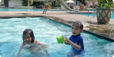 Two young children play in a swimming pool under the sun near a golf course. One child, wearing a blue swimming costume, holds a green bucket and smiles, while the other plays nearby. Poolside and a waterslide are visible in the background.
