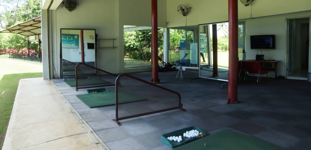 Indoor golf driving range with four practice bays, green mats, and a basket of golf balls in the foreground. Inside, mirrors, a TV, and golf club equipment are visible, with windows overlooking greenery like a proper golf course.