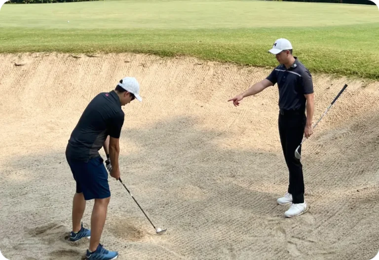 Two men in a golf bunker on the golf course; one prepares to hit a shot with a golf club, while the other, also holding a club, points and appears to give instructions. Both wear caps and typical golf attire.