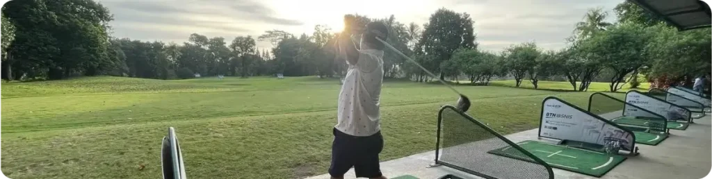 A person swings a golf club at a driving range on a scenic golf course during sunset, with green grass, trees in the background, and several practice bays visible on the right-hand side.