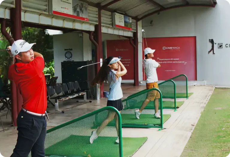 Three people are practising their golf swings with golf clubs at an outdoor driving range, each in separate bays. They are mid-swing, facing the green of the golf course, with equipment and benches visible behind them.