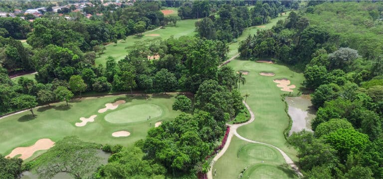 New course - aerial view of a lush green golf course surrounded by dense trees, featuring several fairways, sand bunkers, putting greens, and water hazards, with a few distant golf club buildings in the background.