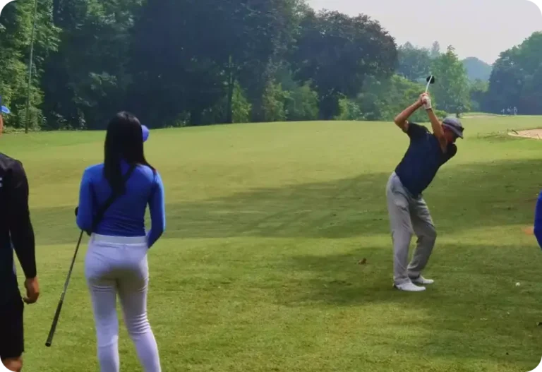 A man is swinging a golf club on a lush golf course while two people, one in blue and one in black, watch nearby. Trees and greenery surround the area under a bright sky.